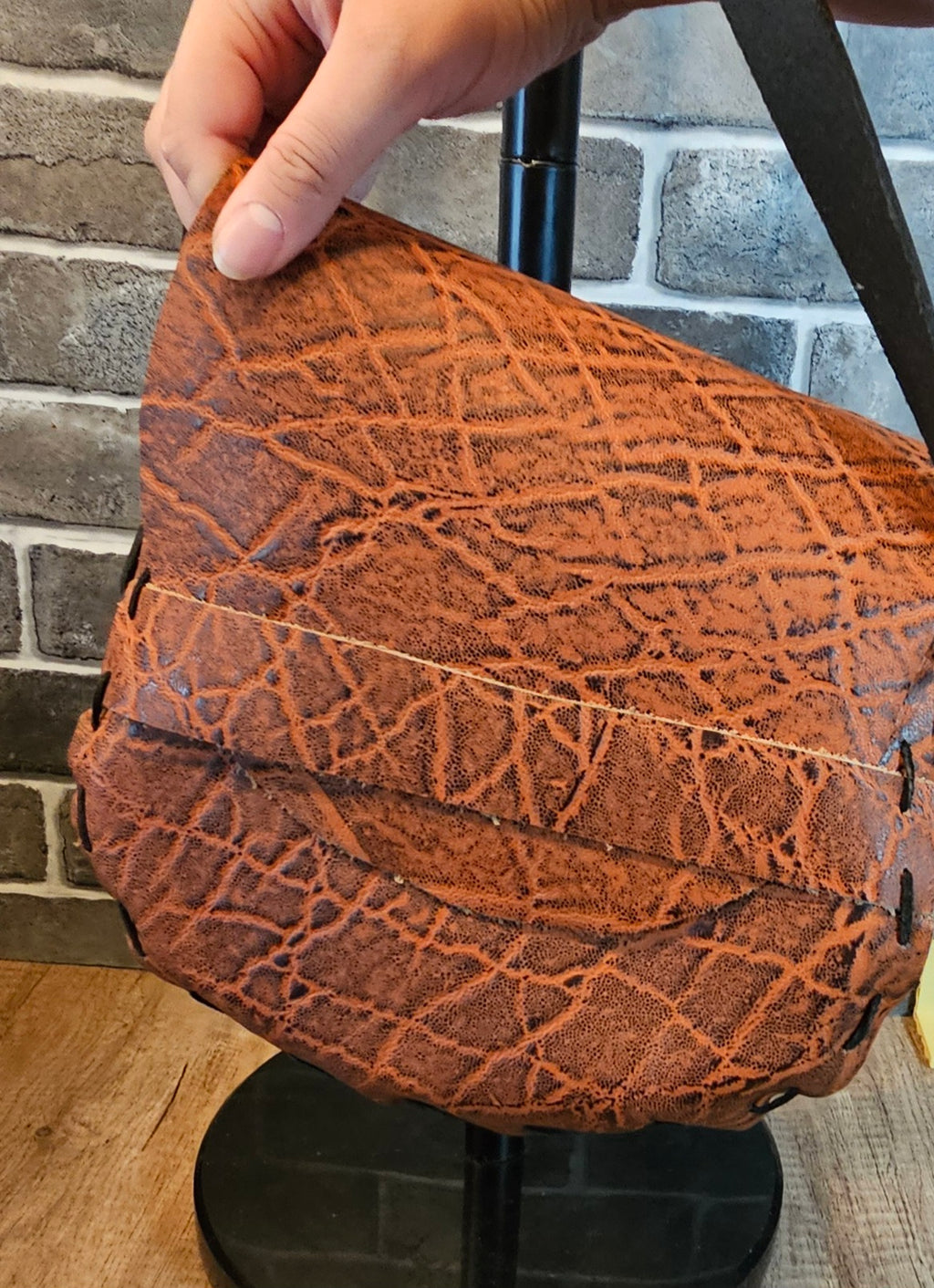 A close-up of a brown textured leather shoulder bag with a distinctive cracked pattern, held by a hand against a gray brick wall background.